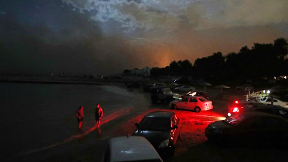 People stand in the sea near to cars parked on the beach to protect them from the fire in the town of Mati.