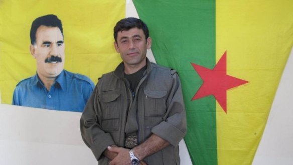 Commander Badr Khan standing under a flag featuring the image of Abdullah Ocalan, the imprisoned PKK leader.