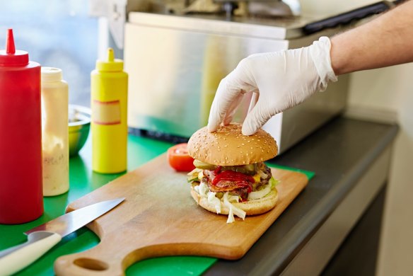 Cropped shot of a white latex gloved hand carefully putting the top bun down on a burger that's been freshly prepared in a kitchen.
Shutterstock image downloaded under the Good Food team account (contact syndication for reuse permissions).